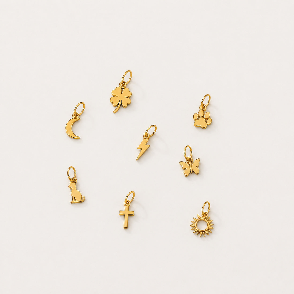 Seven small gold charms with different designs, including a four-leaf clover, crescent moon, paw print, lightning bolt, butterfly, cat silhouette, cross, and sun, arranged on a plain off-white background.