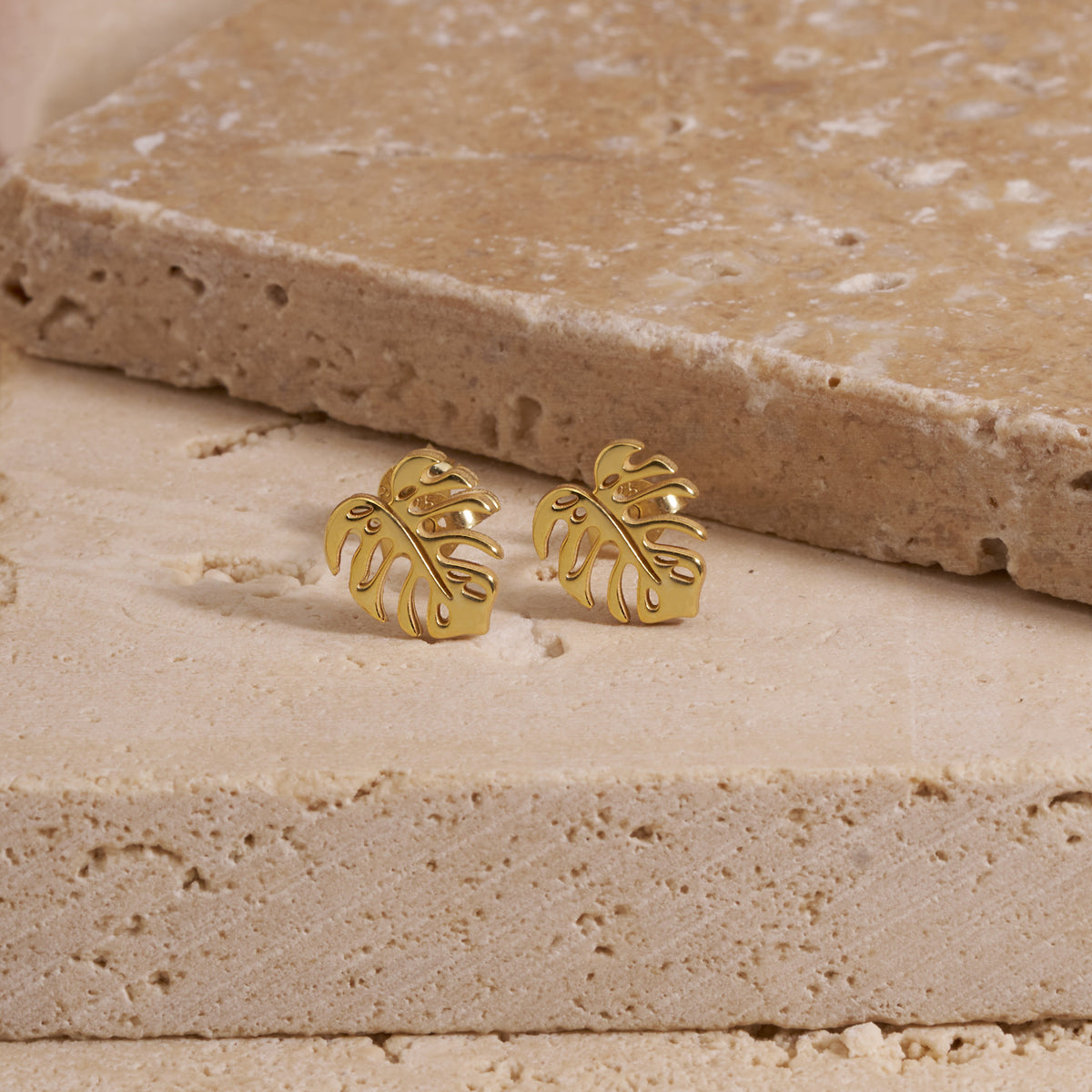 Two gold monstera leaf stud earrings resting on the fingers of a hand