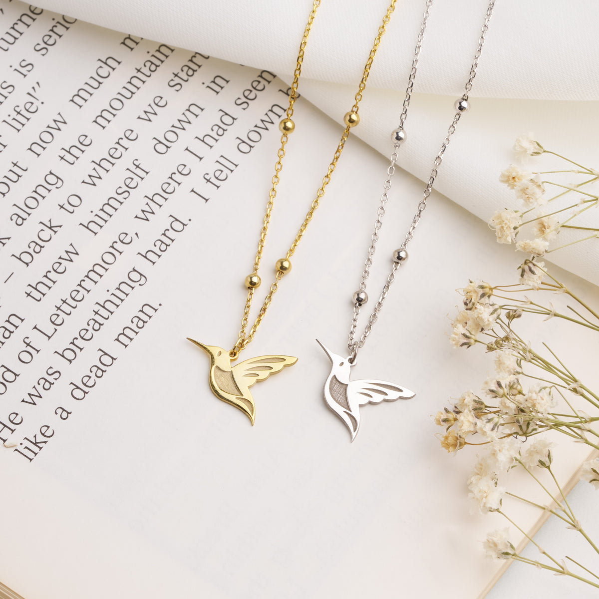 Gold and silver hummingbird necklaces on beaded satellite chains, laid on an open book