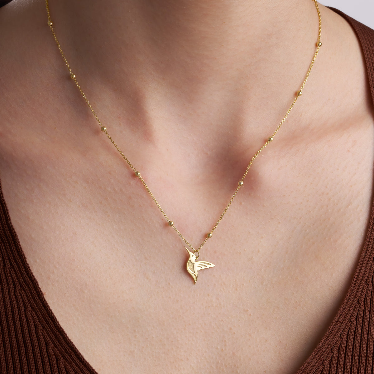 Gold hummingbird necklace on a beaded satellite chain, worn with a brown V-neck top