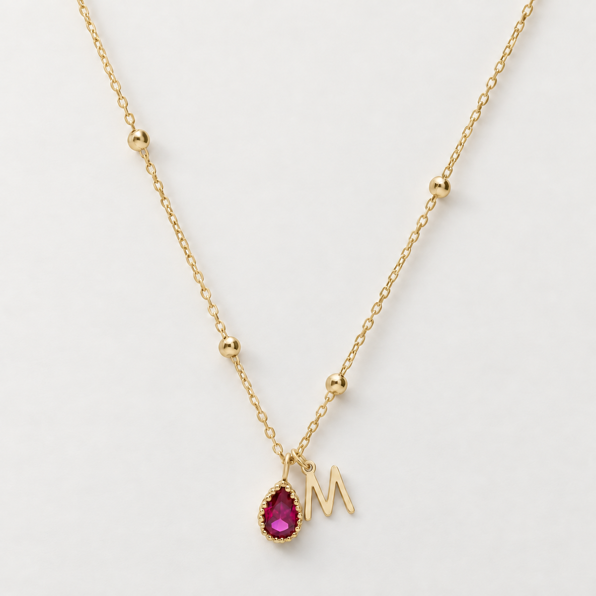 Gold beaded chain necklace with a pink pear-shaped birthstone charm and an M initial charm