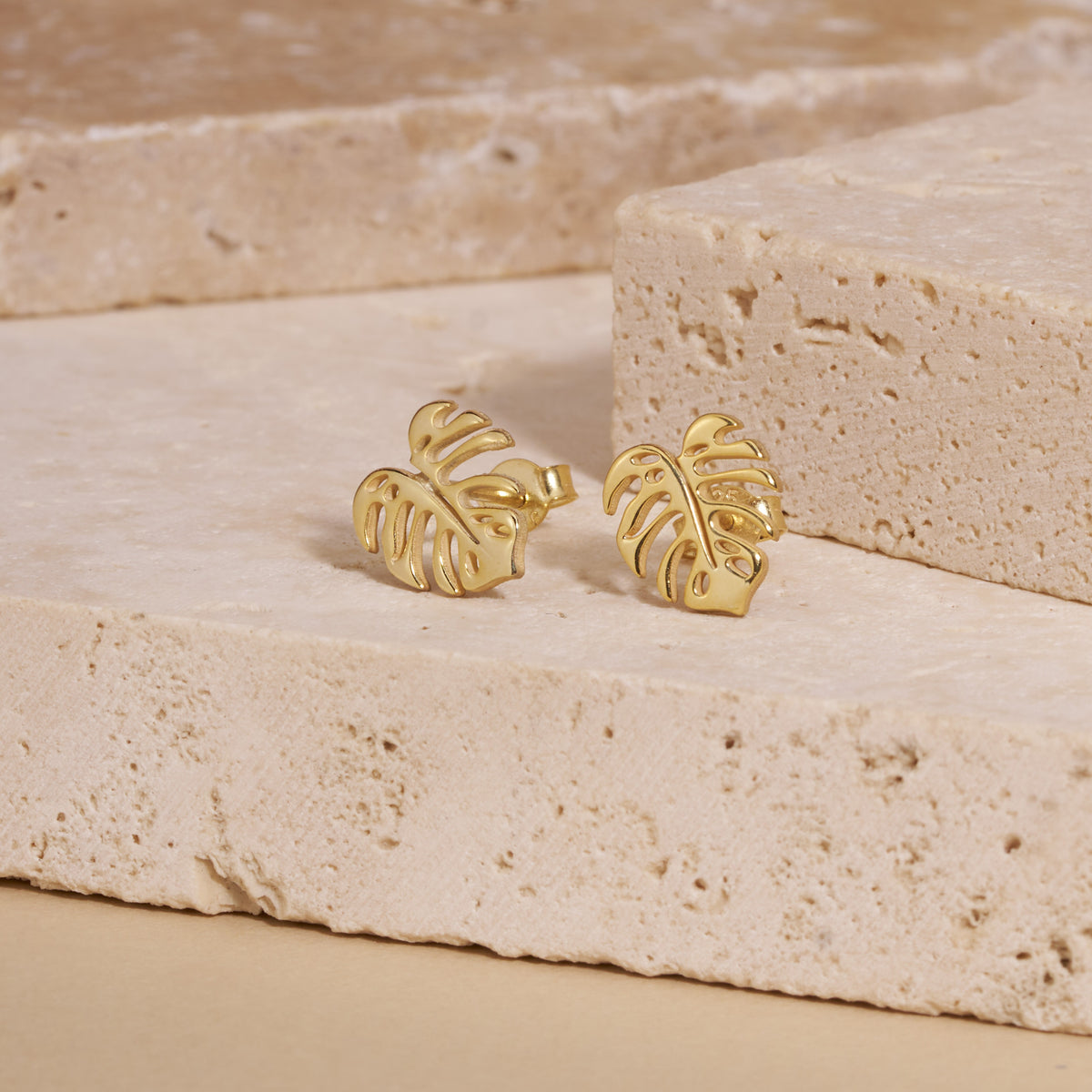 Gold monstera leaf stud earrings at an angle on travertine, showing the post and back