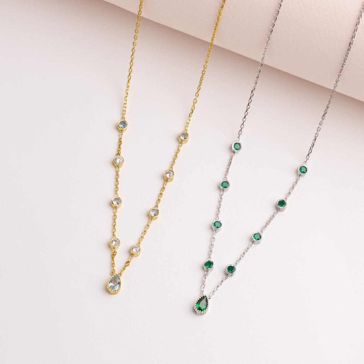 Gold and silver birthstone necklaces laid flat, one with clear and light blue stones, one with green stones