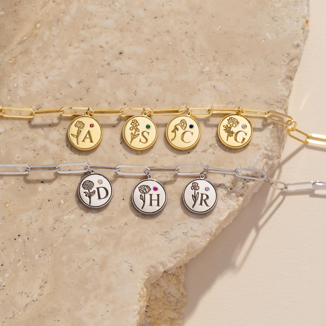 Two gold and silver bracelets with circular charms featuring initials and floral designs on a beige stone surface.