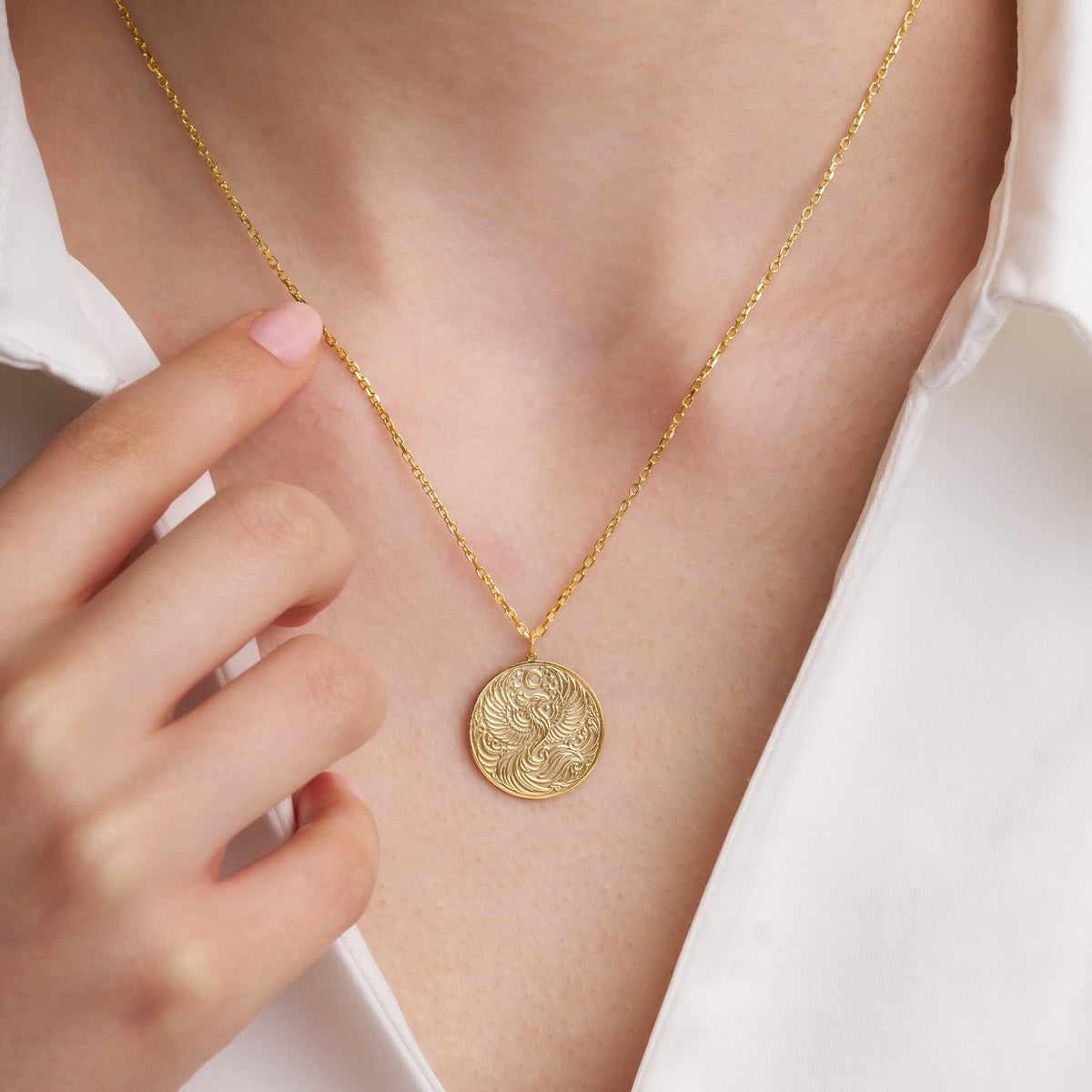 Hand holding the chain of a gold phoenix disc necklace worn on the neck