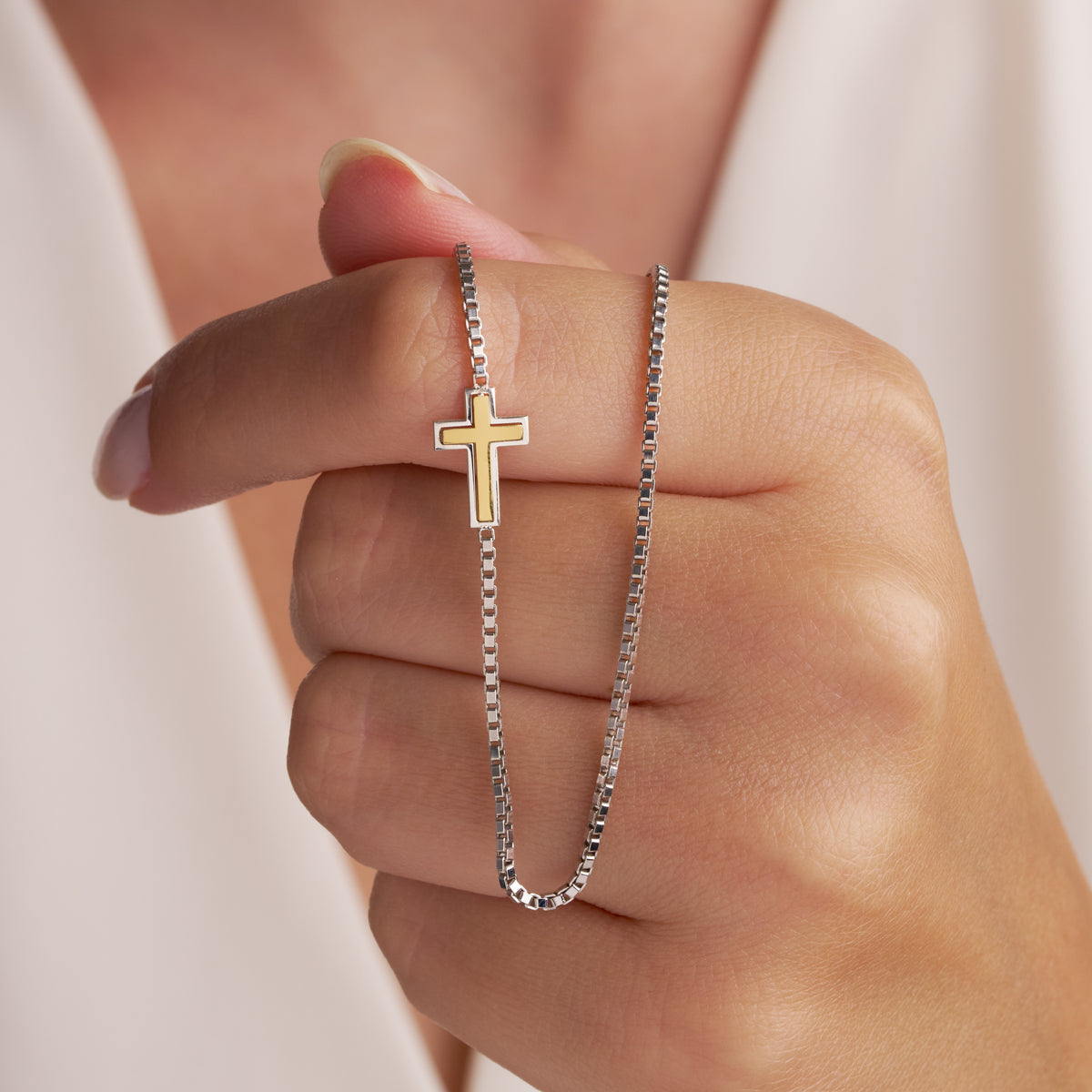 Two-tone sideways cross necklace held over fingers: gold-tone cross in a polished silver frame, set into a box chain